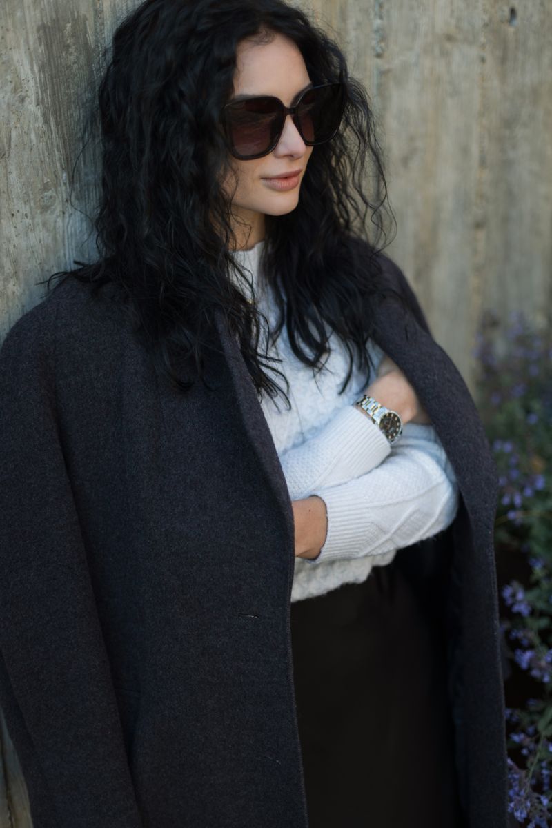 Woman with a dark coat draped over shoulders and oversized sunglasses by a cement wall