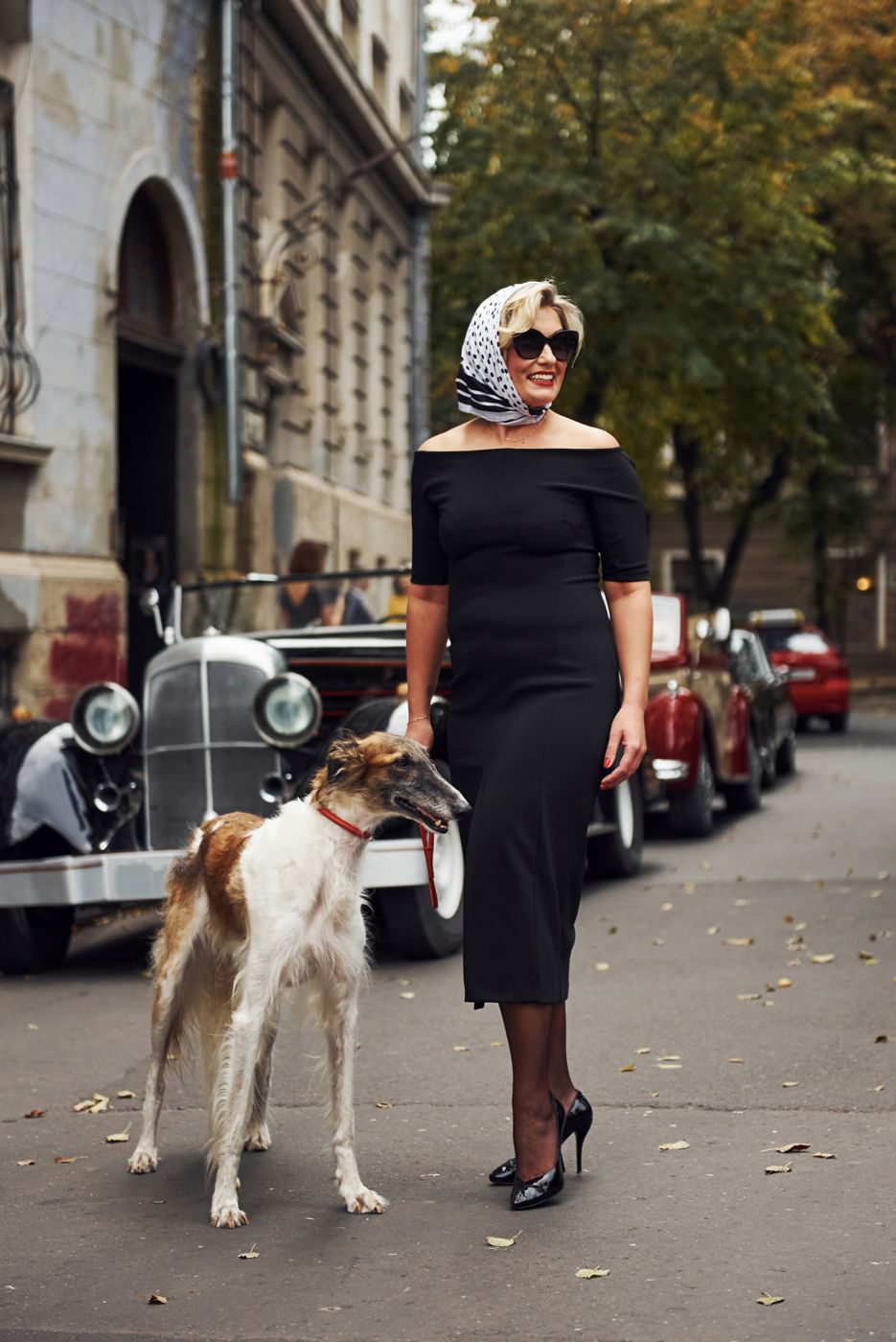 Woman in a black dress with a small dog beside a vintage car