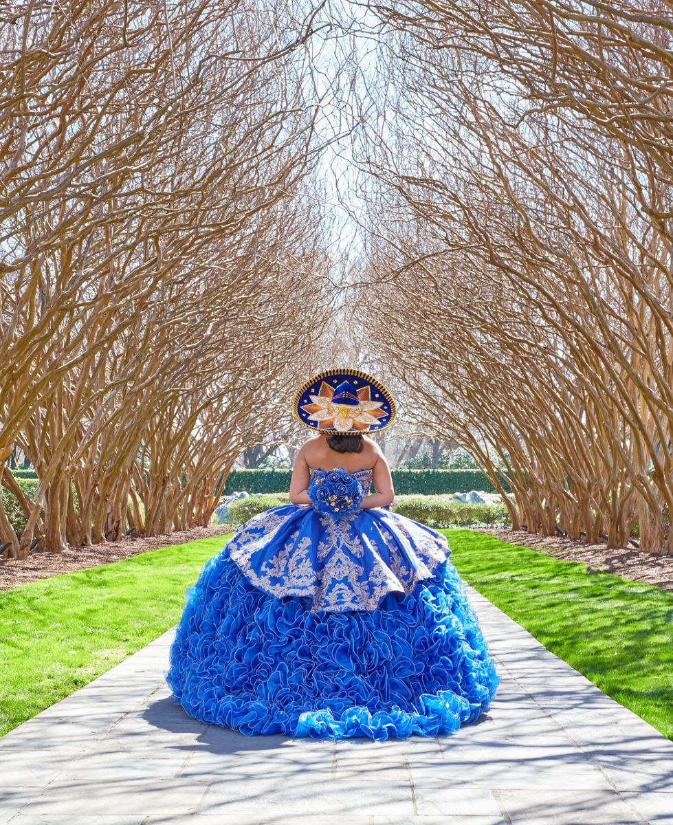 Quinceañera in a royal blue ball gown and charro hat in a tree-lined path