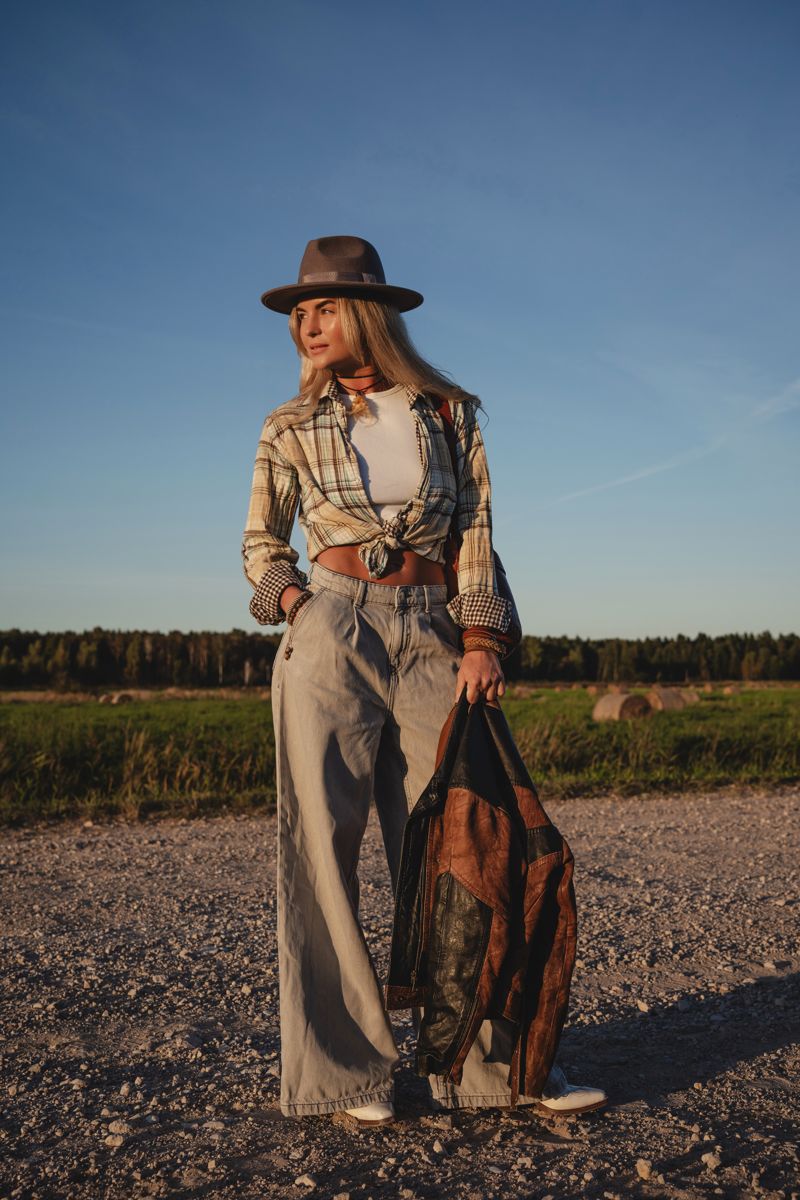 Woman in plaid, hat and wide-leg denim walking a gravel road at golden hour